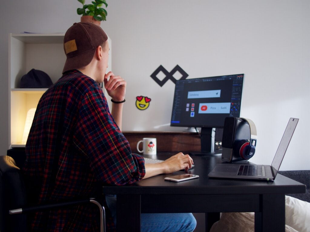 A man wearing a hat is using a computer beside a laptop over a black table.