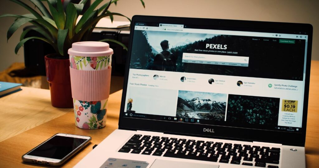 A Dell laptop with a tumbler and a phone on its side displays the pexels website homepage on its screen.