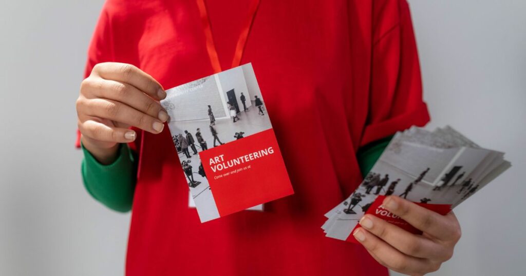 A person wearing a red shirt and ID lace with a green sweater is holding a flyer for art volunteering.