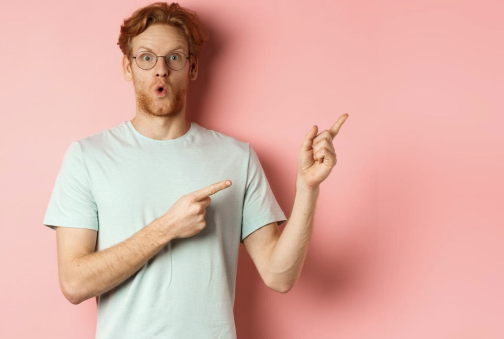 A man on a pink background who looks impressed is pointing both of his index fingers in the air.