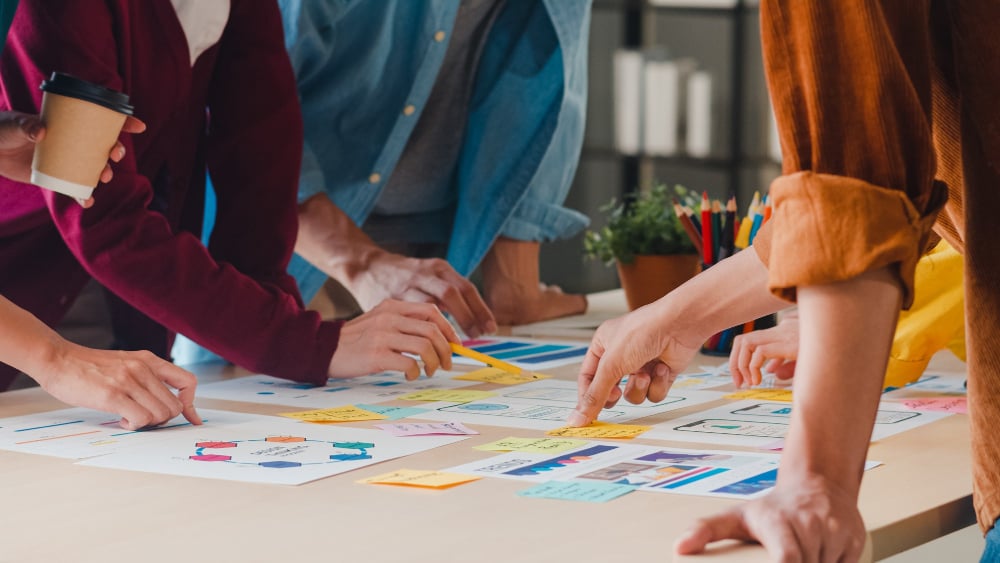 team members are gathered around pointing different papers who are probably brainstorming creative ideas on their design project