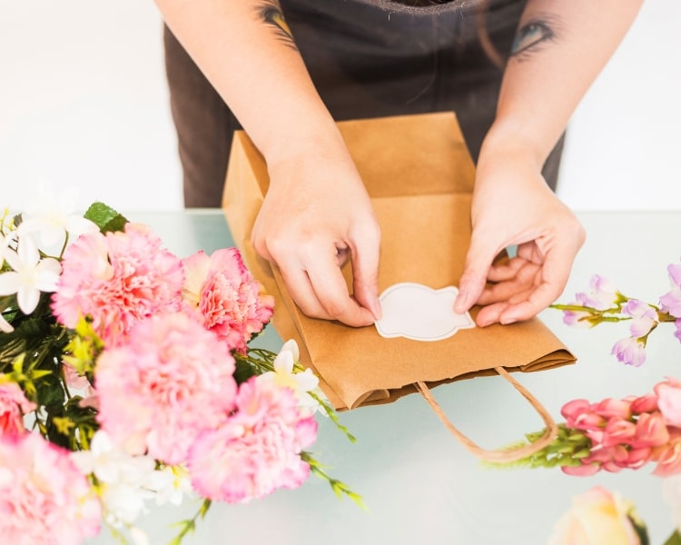 A person with eye tattoos on each forearm sticks a sticker in a store paper bag surrounded by flowers.