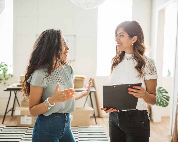 Two women are discussing something in a room with plenty of boxes, and one of the women is holding a black checklist.