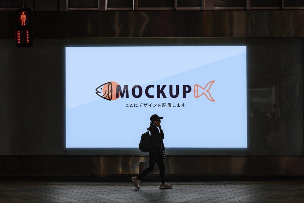 A lady wearing a facemask is passing by a billboard that displays a logo design of a fish logo mockup.