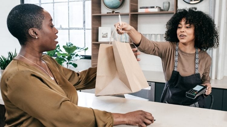 A woman is handing out a no-brand paper bag package on the counter to another woman.