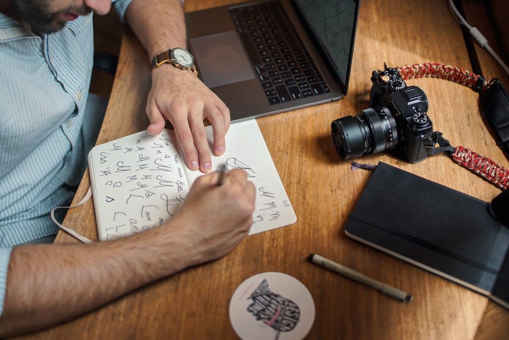 A bearded man wearing a watch is sketching different style for the letter M on a notebook to create a quality logo