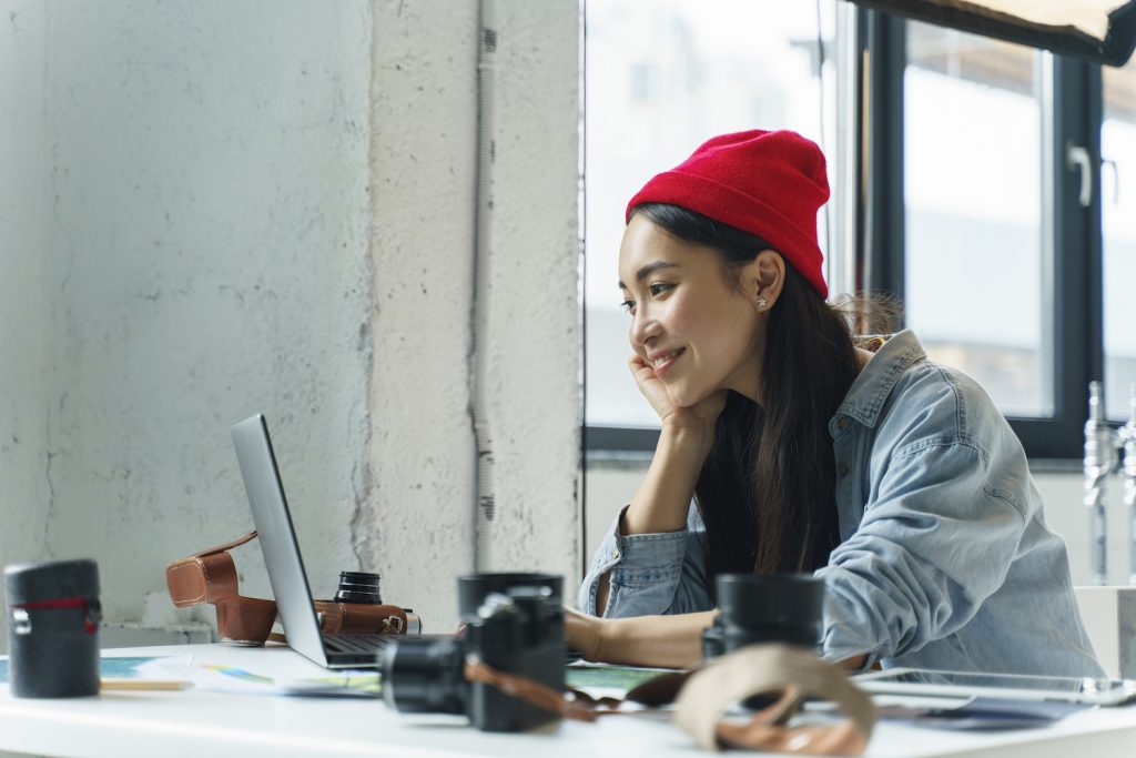 A smiling girl wearing a red bonnet is using a silver laptop on a table. 