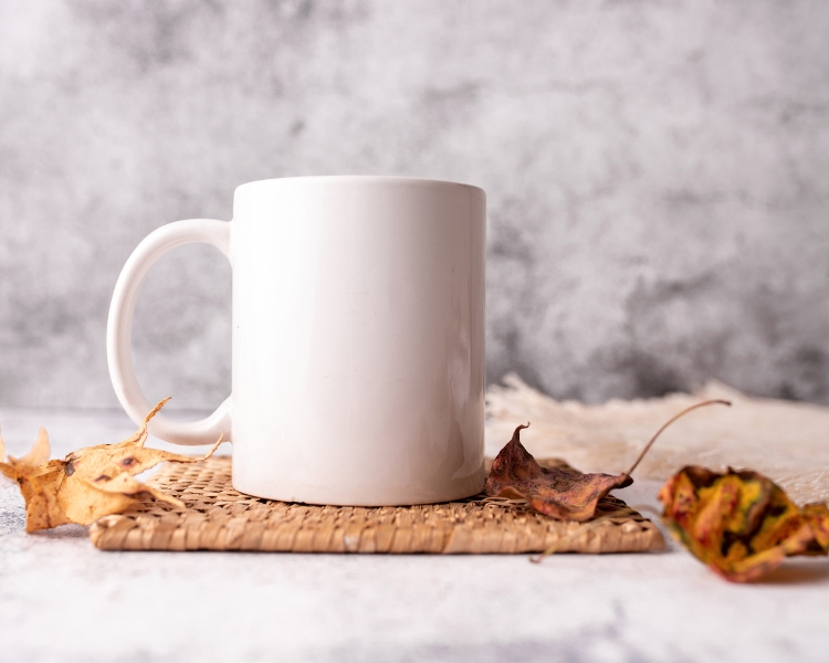 A plain white mug over a woven saucer against a blurry background.