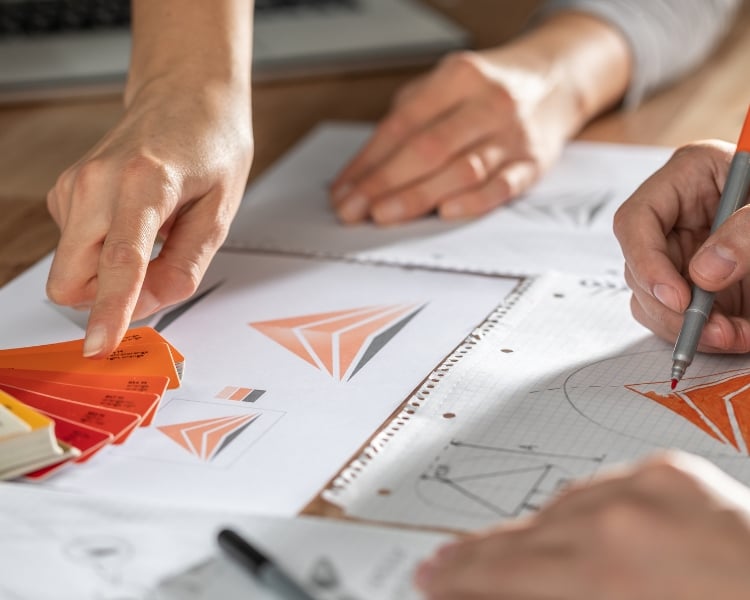 A person is sketching an orange DIY Logo on a physical draft sketch in a paper.