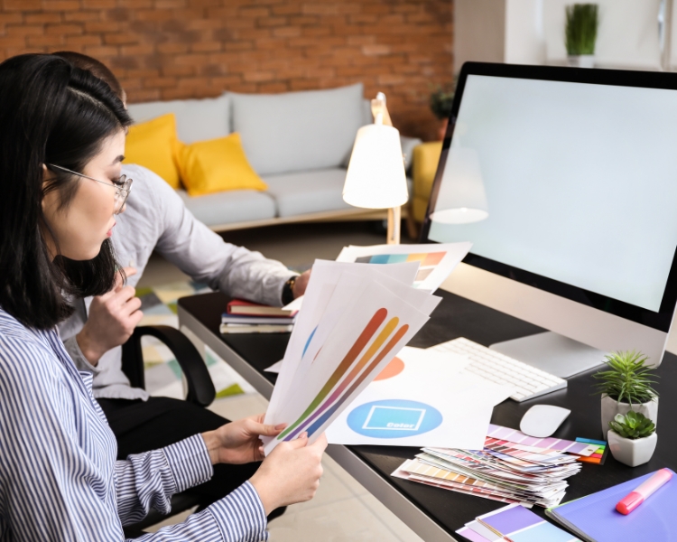 designer comparing colors printed on a paper in front of a monitor and other design materials