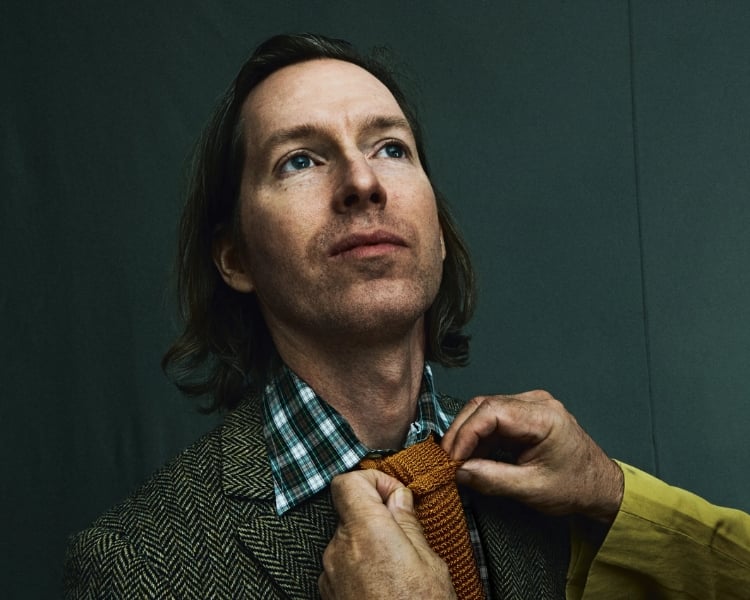 A photo capturing filmmaker Wes Anderson looking upwards while someone is tying a necktie around his neck.