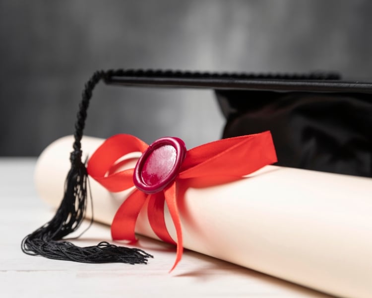 A rolled diploma sealed using a red ribbon with a black graduation cap behind.