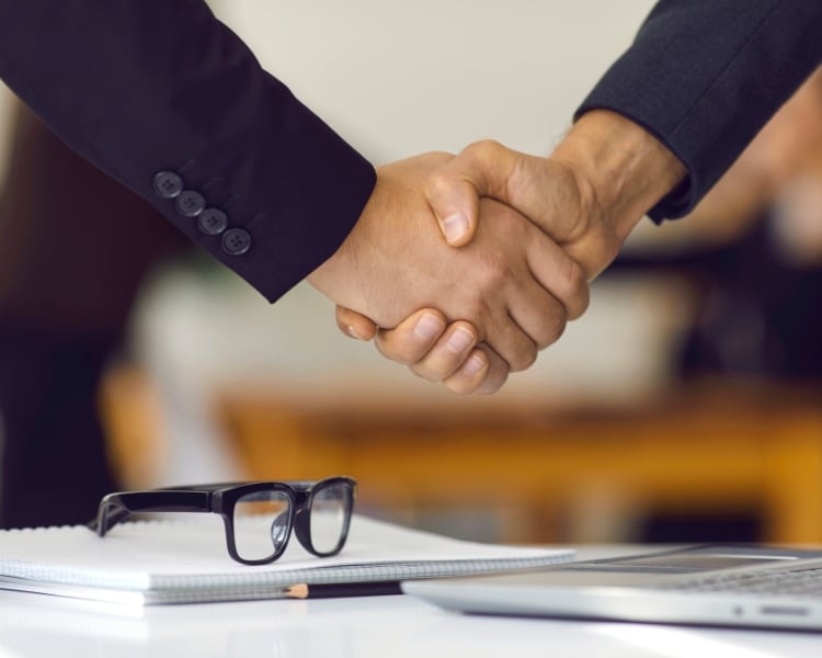 photo of two people secured a deal through shake hands, which camera focuses on the hands only and a glasses and notebook on the table