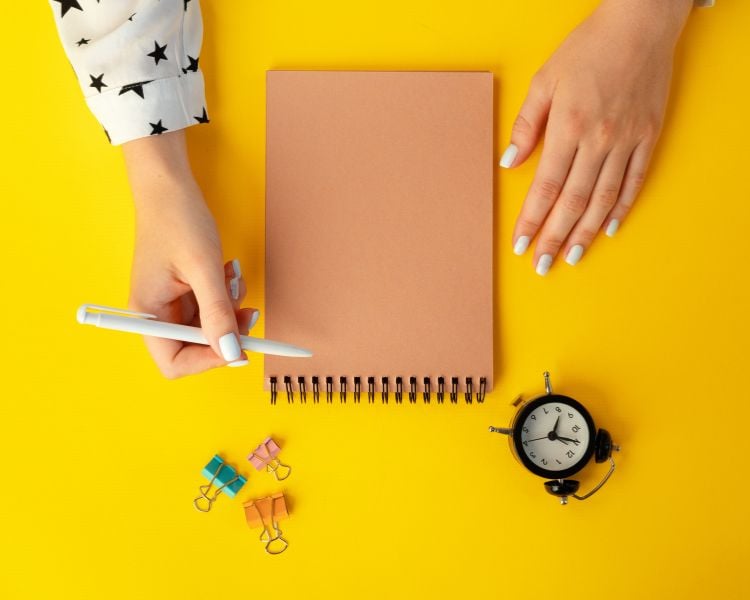 A hand, notepad, white pen, clips, clock, in a yellow background